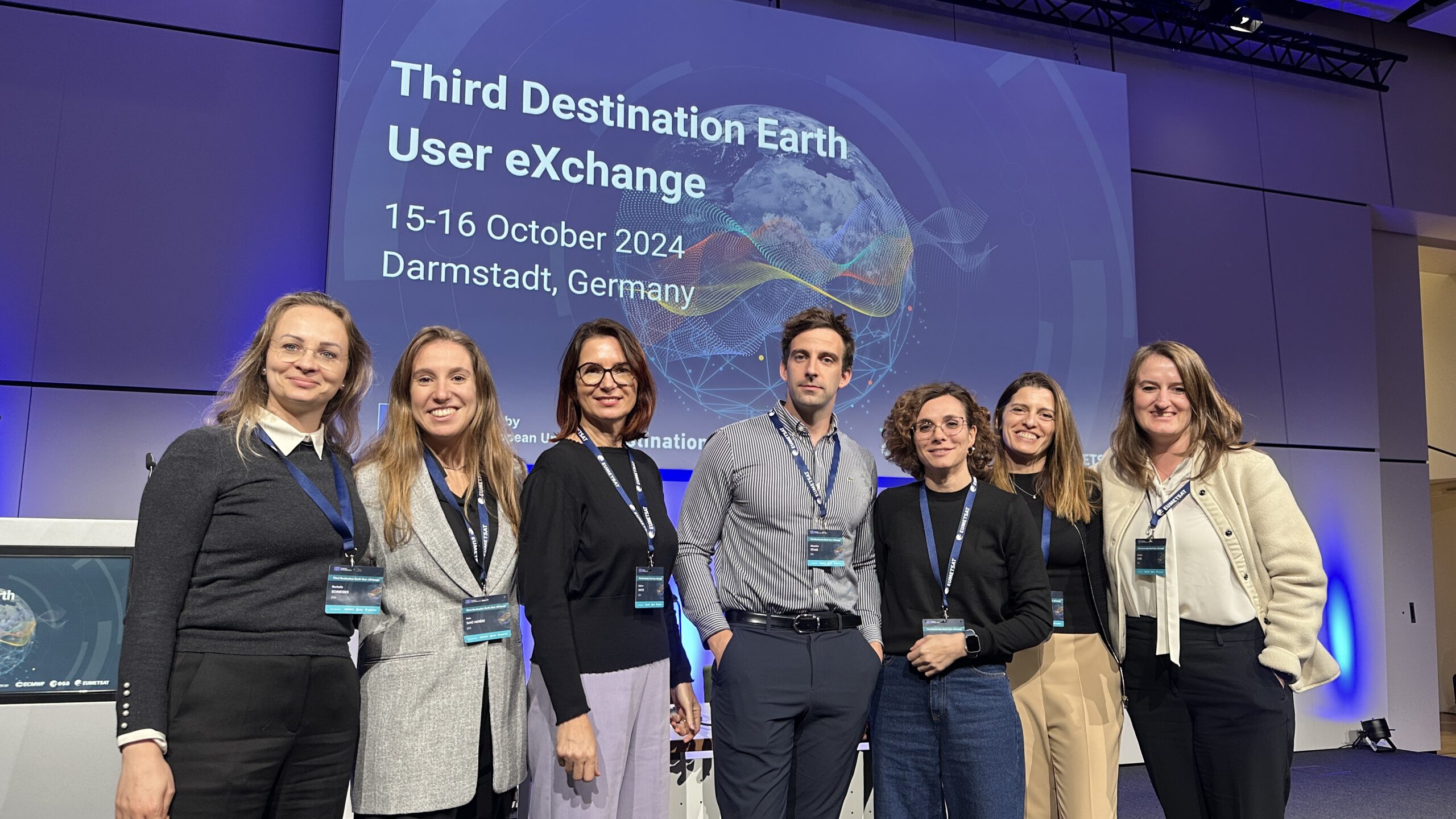 Group picture of six people. They are smiling. In the background, a projection reads "Third Destination Earth User Exchange, 15-16 October 2024, Darmstadt, Germany"